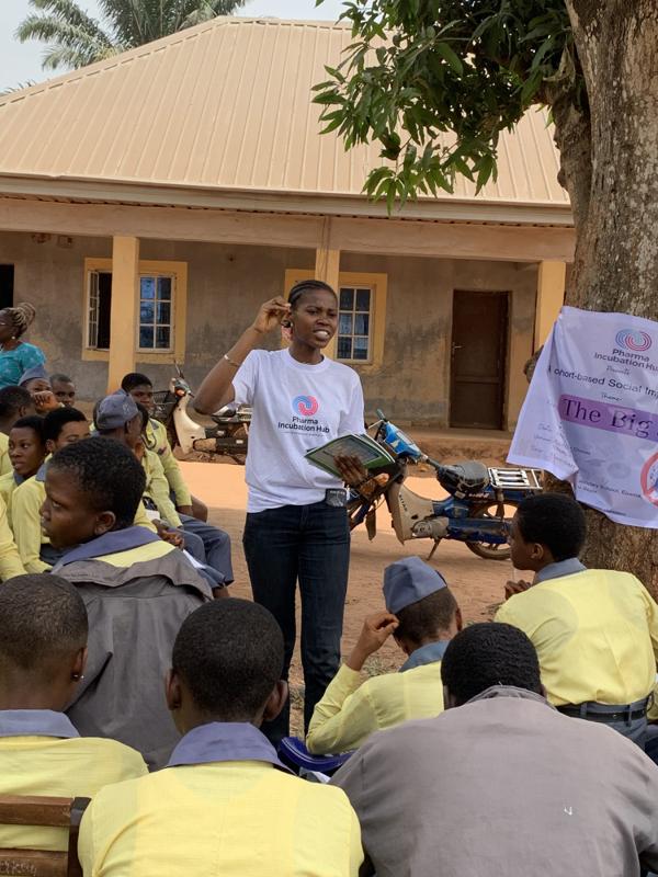 A fellow holds an open workbook and gestures while addressing students seated in a school courtyard
