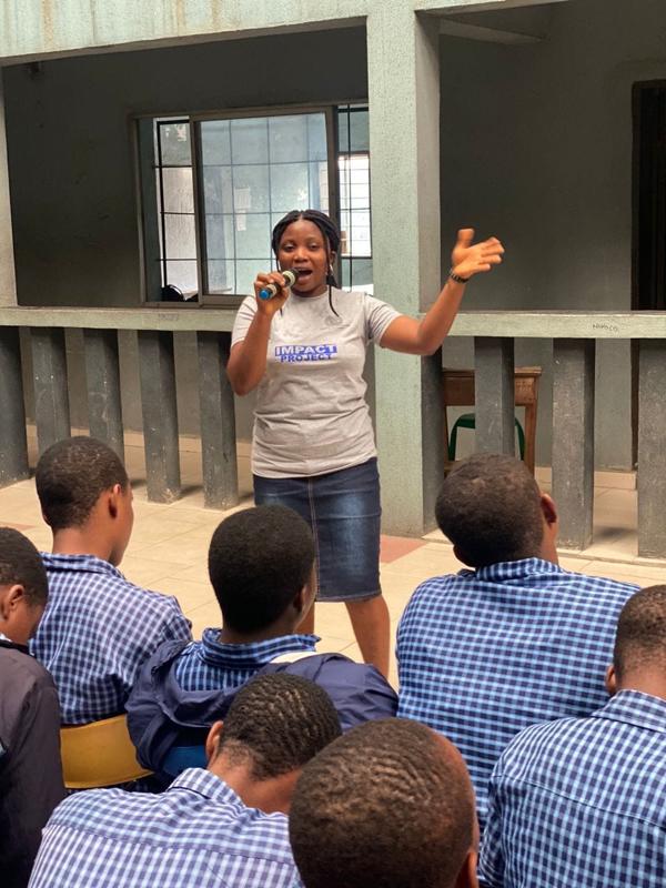 A fellow in an Impact Project shirt speaks into a microphone in front of rows of seated students